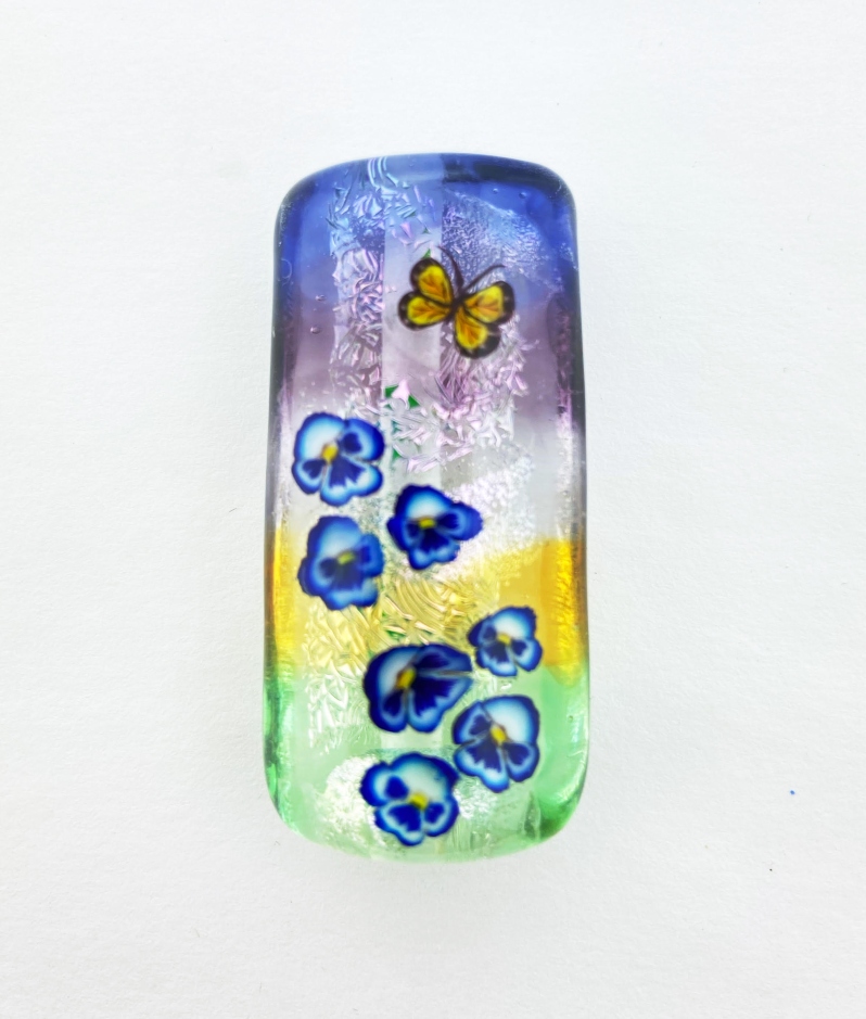 Product Description: 2022 Commememortive Beadby Stephanie White</br></br>Stephanie began lampworking in 2001 and quickly fell in love with the process. Working out of her family owned bead store (Beads by Design) just north of Atlanta Georgia she has spent the last couple of years focusing on her murrine making skills and the 2022 commemorative bead captures her style and work beautifully.</br></br>This commemorative bead starts out with a core that is wrapped with dichroic glass and layered with bands of transparent colored glass loosely inspired by the colors that can be seen during a sunset. The glass is then pressed into a kalera shape and decorated with pansy flowers and a butterfly. Since ancient times the pansy has been a symbol of remembrance while the butterfly symbolizes transformation and growth. </br></br>This bead measures 44mm x 21mm and is 14mm wide . Made on a 3/32 mandrel giving it a 2.5mm hole . ( 1.74 inches x 0.83 inches)</br> 2022 Commemorative Bead