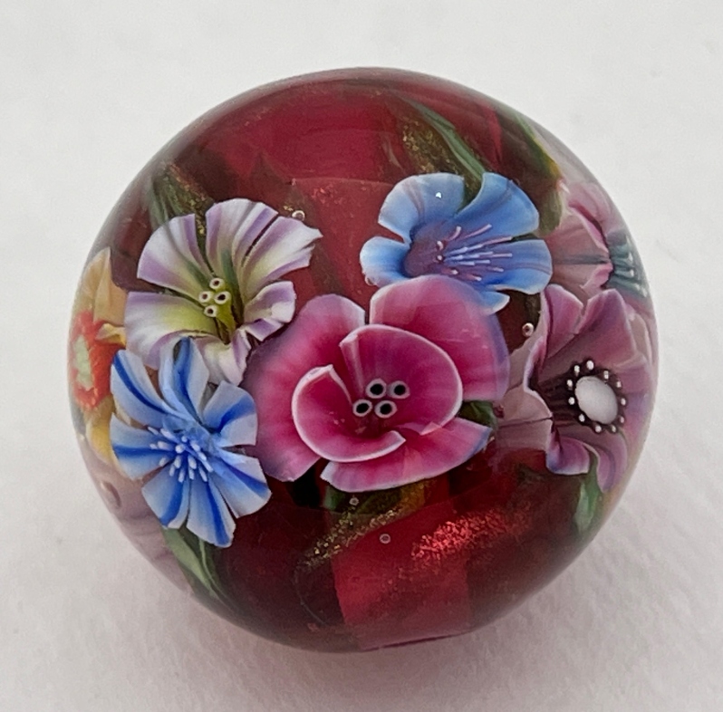 Product Description: Carol Ann has created the 2023 Commemorative bead in the paper weight style. The ruby rich background with goldstone stringer makes a beautiful background for 10 handmade murrini. The bead measures 1” in diameter. </br></br>About the Artist:Canadian artist Carol Ann Savage currently lives in Toronto, Ontario where she is active in the community as a professional musician and a soft glass artisan. She has taught the flute for many years at Upper Canada College and will be teaching glass art across North America in the coming year. Carol Ann is also a Director at Large for the International Society of Glass Beadmakers. Her work can be found in the Corning Museum of Glass gift shop and other galleries around Ontario. Carol Ann was the first prize recipient of the 2022 "Your Bead for Venice" competition.</br></br>Limited Edition -$110.00</br></br>Custom Comemorative Bead Box included.</br> 2023 Commemorative Bead